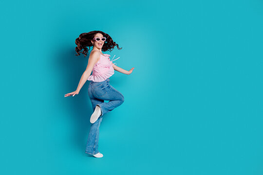 Joyful young woman in pink top and sunglasses jumping against bright turquoise background wearing jeans and white sneakers for playful summer fashion and carefree lifestyle portrait