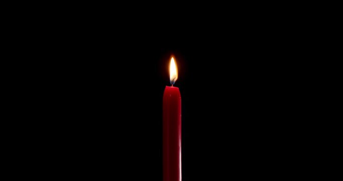 Brightly lit red candle burning with a warm glow in the dark. A striking wax candle that contrasts with a completely black background. This classic candle represents memory and celebration.