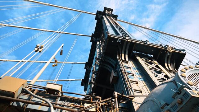 Manhattan Bridge tower and suspension cables from below. Riveted steel beams rise against blue sky streaked with thin clouds.