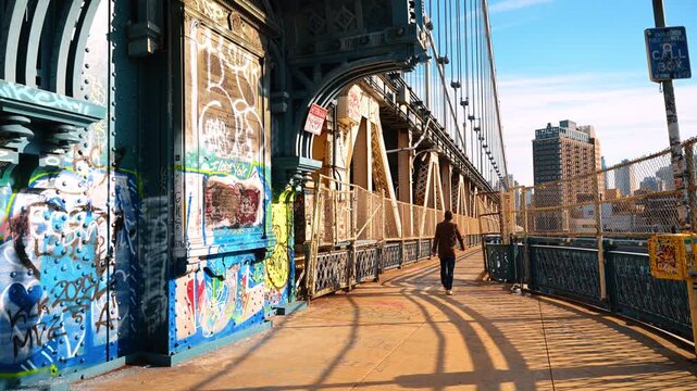 Pedestrian walking along graffiti covered Manhattan Bridge. Late sunlight casts long fence shadows across the walkway beside blue steel arches.