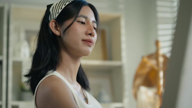 Young Asian woman artist painting on canvas easel with paintbrush in art studio, representing creative professional women designer, fine arts student lifestyle and freelance illustrator