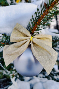 A silver ball hangs from a branch of an evergreen tree covered in snow. The golden ribbon ties the ornament to the branch, adding to the festive mood
