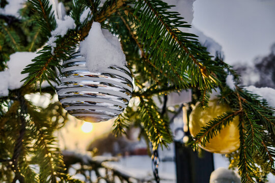 Ornaments hang from a decorated Christmas tree covered in snow. The sun sets in the background, showing a winter evening atmosphere