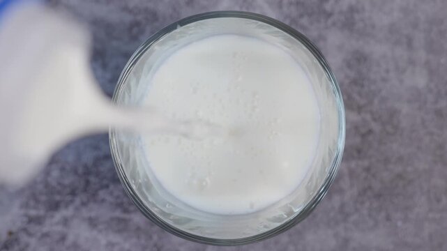 Top view close-up video of fresh white milk being poured into a clear glass, surrounded The creamy white milk fills the glass	