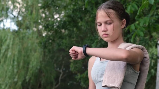 Teen girl checking smart watch tracker during outdoor training in summer park. Concept of activity monitoring device
