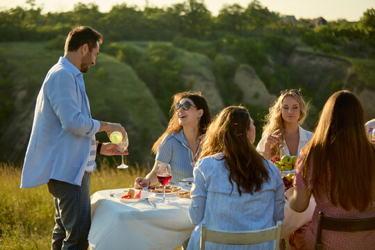 Male host serving drinks to women seated at countryside dinner table. Concept of attentive hospitality, beverage service, premium outdoor entertaining.