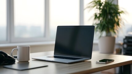 Fototapeta premium A sleek laptop sits open on a wooden desk beside a white mug and smartphone near a bright window