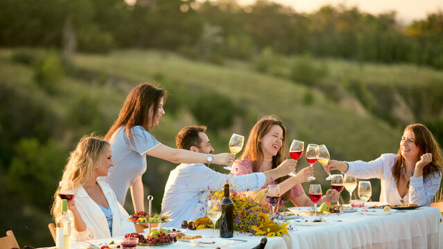 Women reaching across outdoor table to clink wine glasses in celebration. Concept of female community, friendship campaign, celebratory beverage marketing.