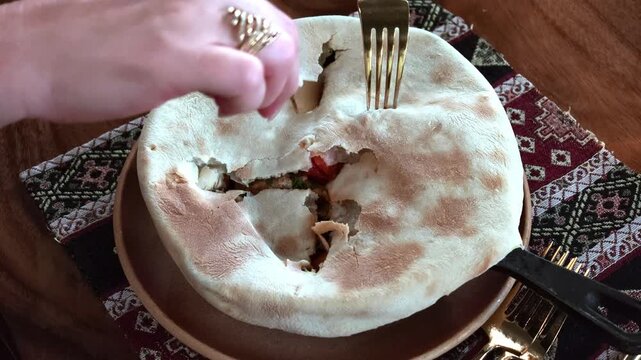Stuffed flatbread with vegetables and meat being sliced beside decorative textile, warm rustic meal with visible hands, jewelry, plate, crust, filling, table setting and rich texture