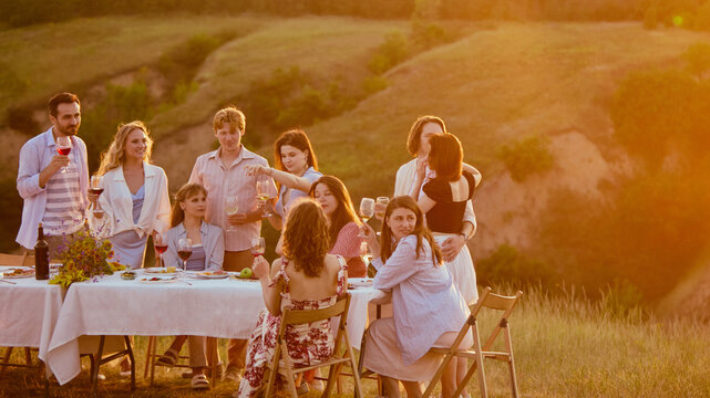 Young friends dancing behind dinner table during glowing countryside sunset. Concept of after-dinner entertainment, summer festival mood, carefree youth lifestyle.
