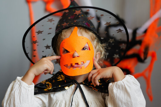 A child smiles while holding a pumpkin mask during Halloween. The background shows decorations in black and orange tones, creating a festive mood