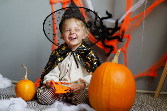 A young girl dressed as a witch smiles while sitting on the floor surrounded by pumpkins and Halloween decorations in a cozy room