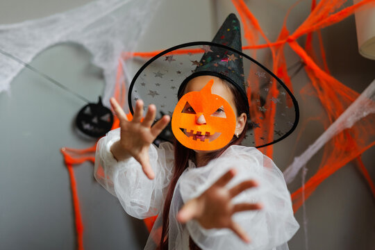 A child plays in a room decorated with Halloween items. The child wears a witch hat and an orange pumpkin mask while reaching out with hands