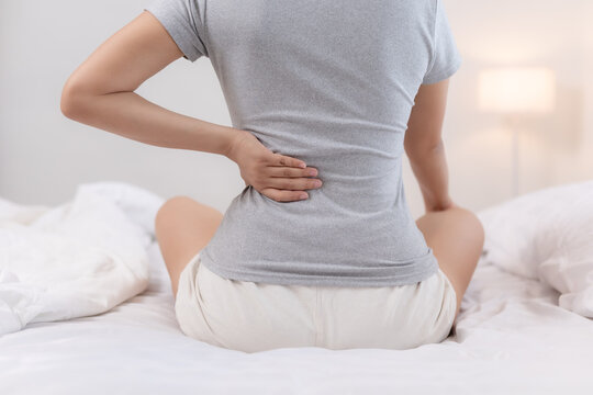 Uncomfortable young woman sitting on bed holding lower back experiencing severe pain suffering from chronic muscle ache during early morning inside quiet cozy bedroom
