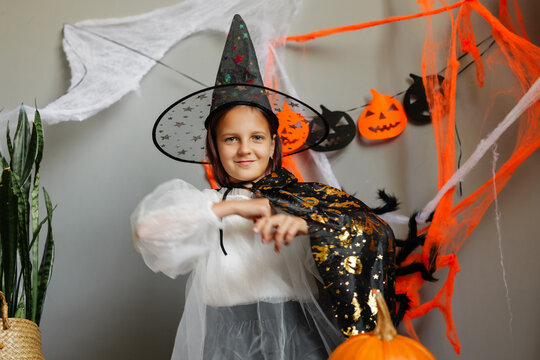 A child is dressed as a witch in a room decorated for Halloween. The child has a pointed hat and a cape while standing near pumpkins