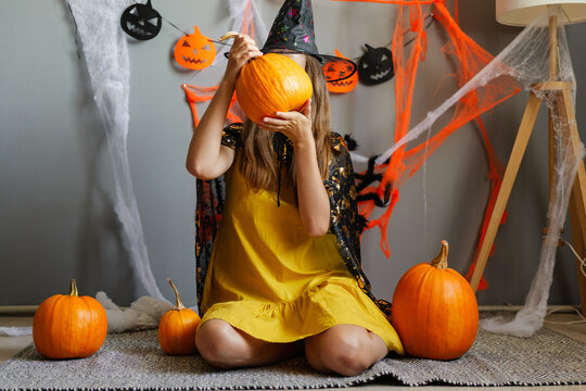A person is dressed in a witch costume and is holding a pumpkin in front of their face. Around them are several pumpkins and Halloween decorations