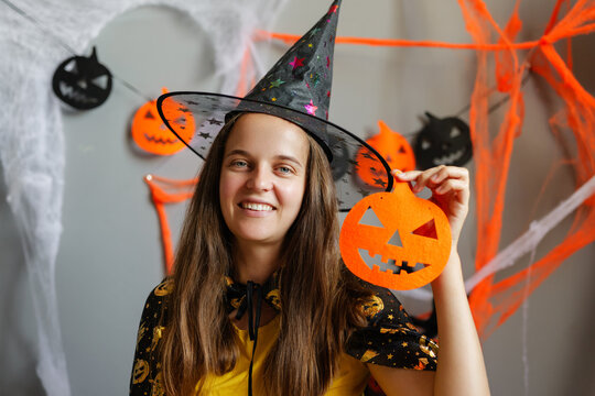 A woman smiles while wearing a witch hat and holding a pumpkin cutout. The background features Halloween decorations like webs and bats
