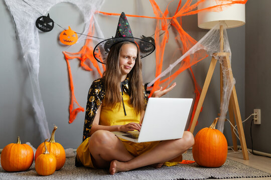 A woman wearing a witch hat and cape sits on the floor with pumpkins around her, using a laptop. Halloween decorations are visible in the background