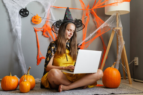 A young woman sits on the floor in a witch costume with a laptop. She is surrounded by pumpkins and Halloween decorations in a cozy room