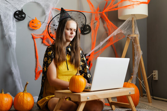 A girl wearing a witch costume works on a laptop in a room decorated for Halloween with orange and black webs and pumpkins around her