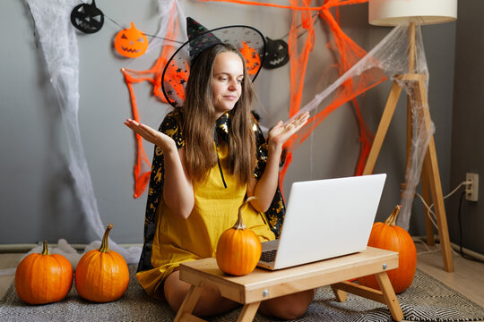 A woman wears a witch costume and sits on the floor with pumpkins. She works on a laptop surrounded by Halloween decorations in a cozy room