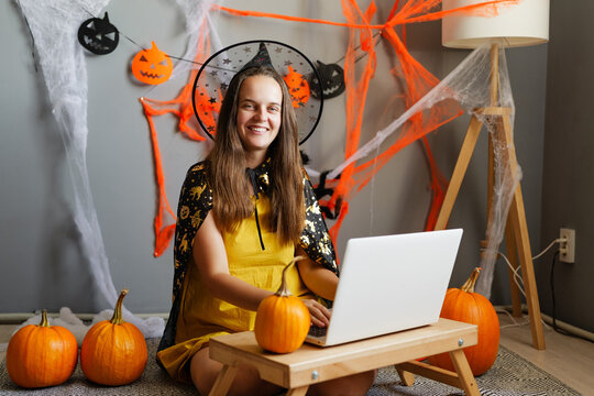 A woman sits at a table with a laptop, smiling while wearing a yellow dress and a black cape. She is surrounded by pumpkins and Halloween decorations