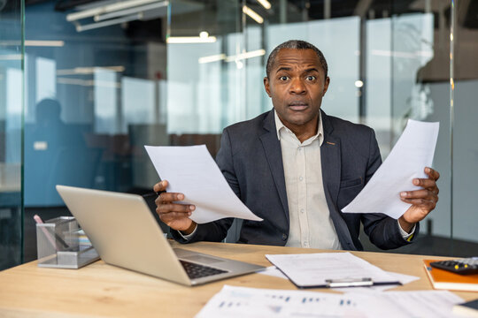 Mature businessman sitting at an office desk, holding paperwork in both hands and reacting with surprise to unexpected information, dealing with stress and confusion in his professional career