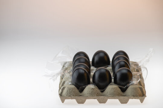 Six black painted eggs in a cardboard carton with plastic packaging film beside it on a warm peach background. Conceptual dark still life, studio photography.