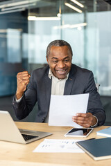 Obraz premium African american businessman celebrating success and achievement, reading a document with an excited expression and clenched fist while sitting at a modern office desk with a laptop and tablet