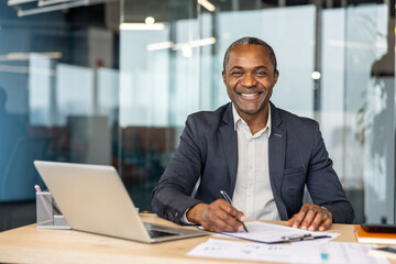 Obraz premium Smiling african businessman writing on documents at his desk, having a laptop and office supplies, portraying success, professionalism, and confidence in a corporate environment