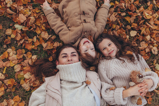 High-angle close-up portrait of happy mother with her children lying on bed of colorful fallen autumn leaves in park. Family happiness, autumn season, childhood joy, motherhood, love and togetherness.