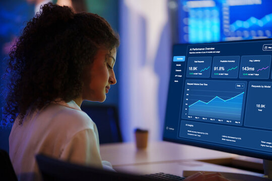 Serious Latin woman monitors an AI cybersecurity dashboard, analyzing network data, system performance, and digital security in a modern office.