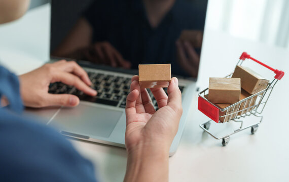 Customer comparing products while shopping online using a laptop with parcel boxes and a miniature cart, representing e-commerce, secure payment and delivery.