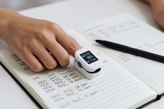 Woman using pulse oximeter on finger to measure blood oxygen level and heart rate. Patient recording vital signs in medical logbook for cardiovascular health monitoring at home.