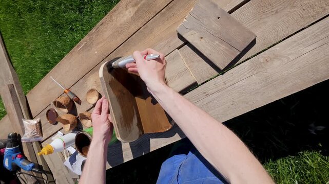 POV recording of a DIY maker staining a wooden bird feeder part using a brush on a rustic workbench. 