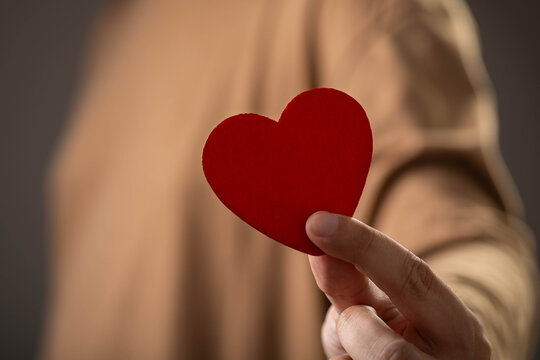 Close-up of a hand holding a red heart symbol representing love, romance, kindness, compassion, relationship, emotional support, charity, healthcare, and human connection with soft background.