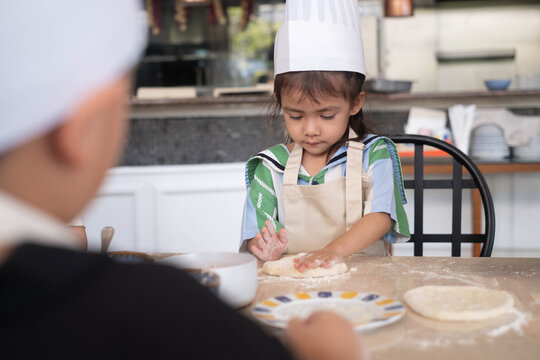 Cute Asian little girl wearing a chef hat and apron, kneading pizza dough during a cooking class with instructor guidance. Hands-on learning, childhood education, baking skills, and family lifestyle.