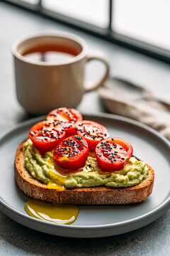 A slice of artisan bread topped with mashed avocado, ripe cherry tomatoes, a sprinkle of black sesame seeds and olive oil.