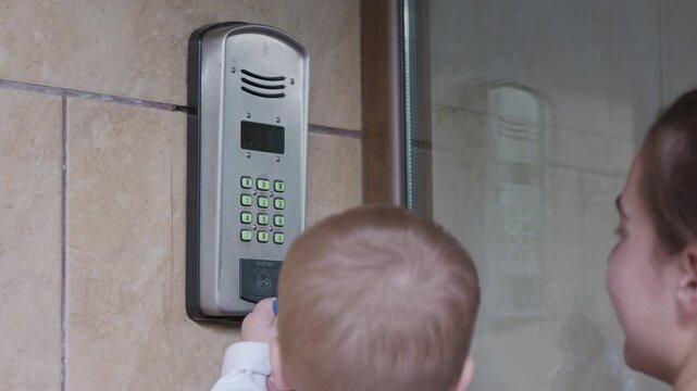 Child interacts with a digital door entry system, pressing buttons on the keypad while an adult stands nearby, both in a residential setting with tiled walls