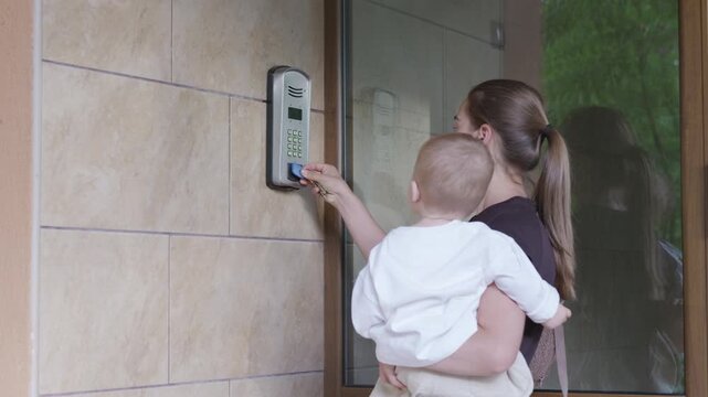 Woman holding baby approaches intercom system at entrance, prepares to press button while standing in front of glass door with tiled wall background