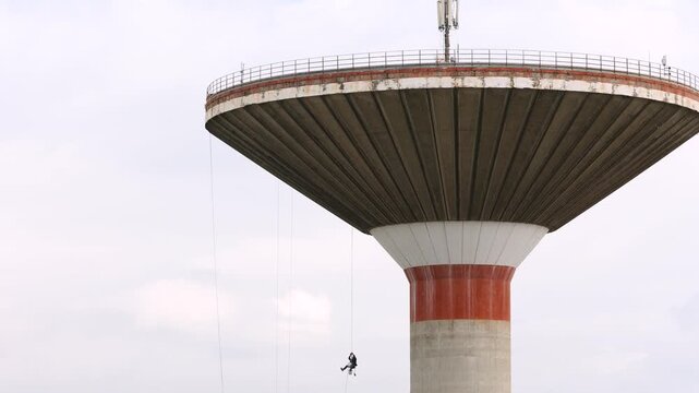 A daring industrial climber suspended on a long rope descends from a huge concrete water tower against a cloudy sky. This specialized maintenance job highlights professional courage and extreme safety