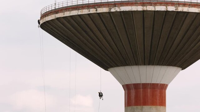 Industrial climbers wearing safety harnesses work meticulously on the curved facade of a towering concrete water structure. This challenging high altitude job demands professional courage and precisio