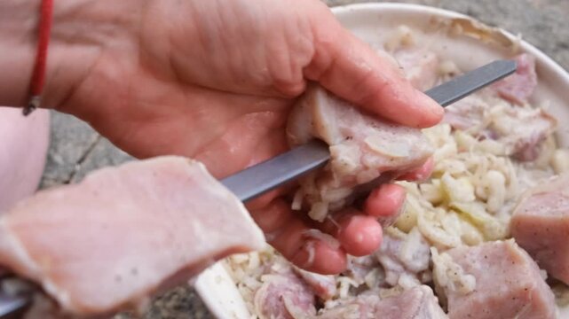 Close-up of hands threading marinated meat onto metal skewers for barbecue, preparing homemade shashlik outdoors with fresh ingredients before grilling over an open fire