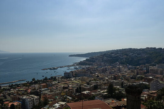 Dense urban architecture borders the calm blue Mediterranean waters along the Naples coastline. This expansive city panorama inspires European travel and highlights majestic Italian heritage.
