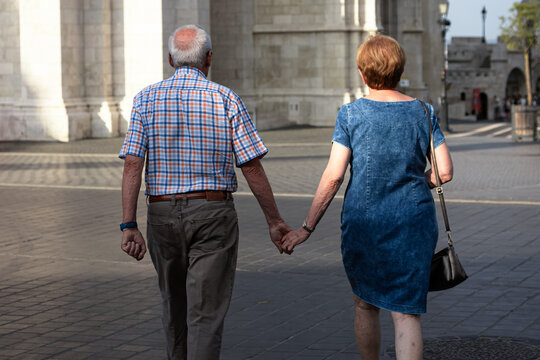 An older couple walks hand in hand across a sunlit paved city square from behind. Such tender moments of togetherness highlight the enduring beauty of lifelong romance and peaceful retirement travel.
