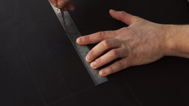 Leatherworker tracing straight lines on black leather using an awl and a metal ruler for a DIY handicraft project
