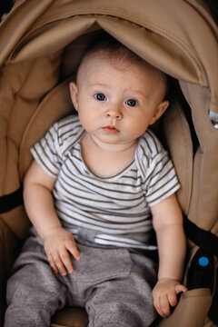 A baby dressed in a striped shirt and gray pants sits in a beige padded stroller, looking forward with a curious and slightly surprised expression. The warm tones and soft lighting create a cozy, cand