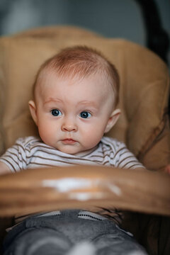 A baby dressed in a striped shirt and gray pants sits in a beige padded stroller, looking forward with a curious and slightly surprised expression. The warm tones and soft lighting create a cozy, cand