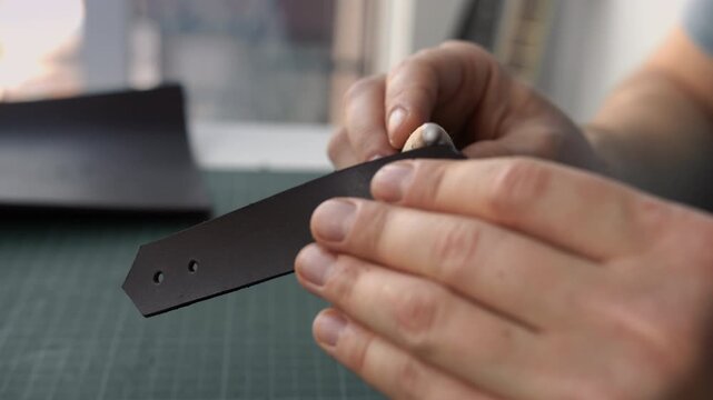 Craftsman burnishing the edge of a handmade brown leather strap using a wooden tool in a workshop
