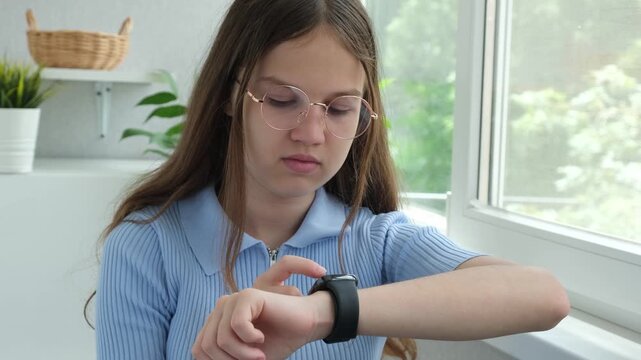 Focused teenage girl in glasses clicks on screen of smartwatch. Sending message by using smart watch. Concept of technology, communication and digital devices